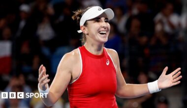 Belinda Bencic, in red tennis top and white visor, celebrates with a smile and arms spread wide, with crowd visible behind her
