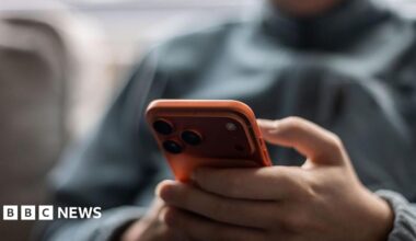 A young person in a grey tracksuit jacket holds a red iPhone.