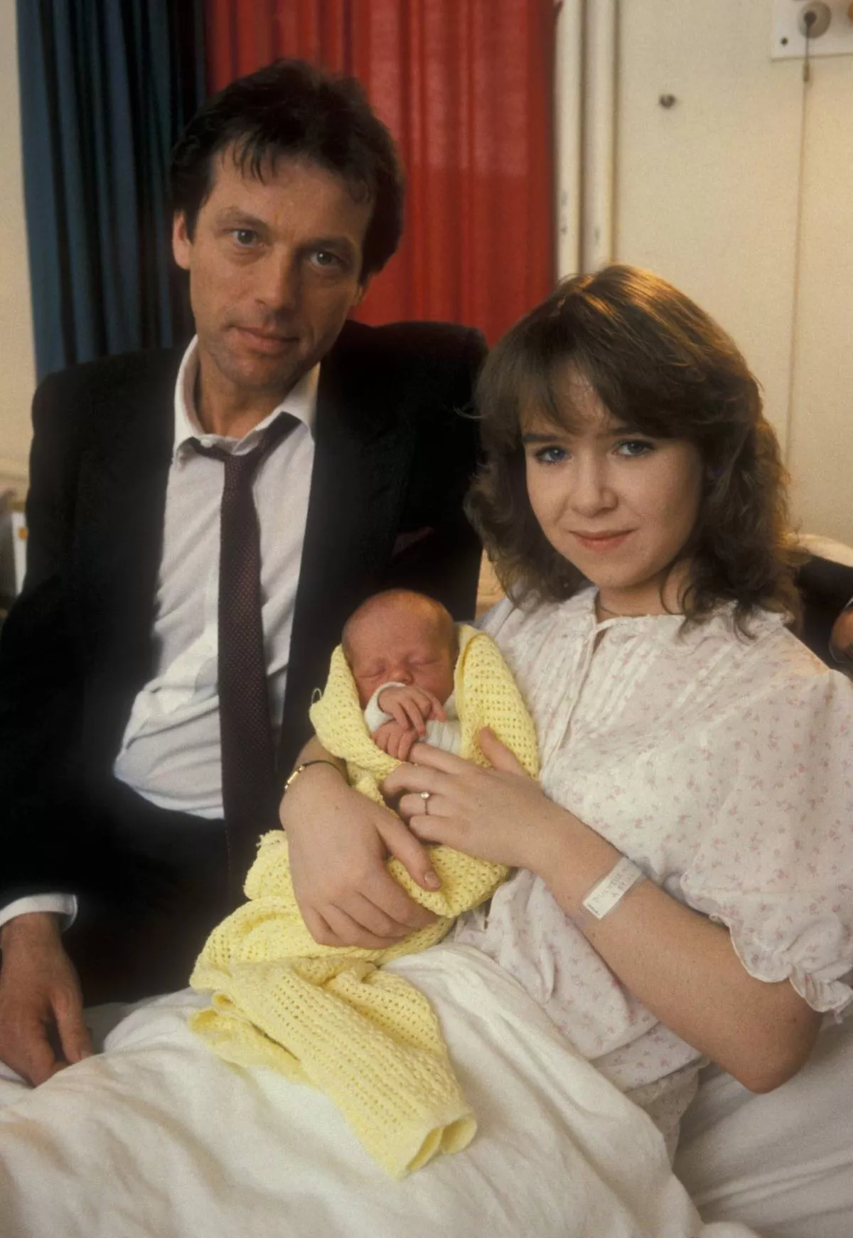 A man and a woman are seated together, with the man dressed formally in a black suit and tie. They are holding a newborn baby, who is wrapped in a light-colored blanket, in their arms. The background features a red curtain and a white wall.