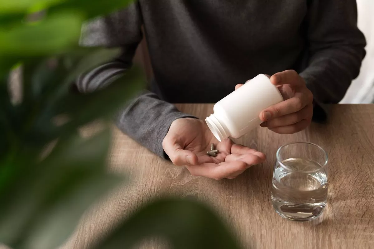Adult man pouring supplement pills from a white bottle into his hand with a glass of water