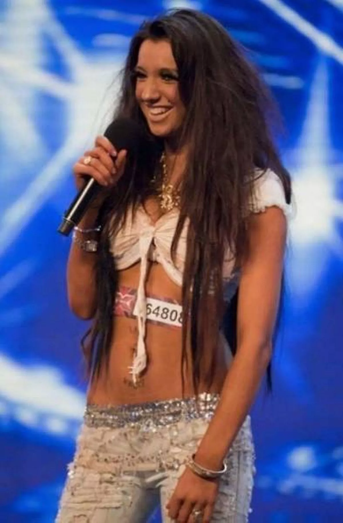 An individual with long, flowing hair stands on a stage, holding a microphone and delivering a speech while a blue background adorned with white stars serves as the backdrop.