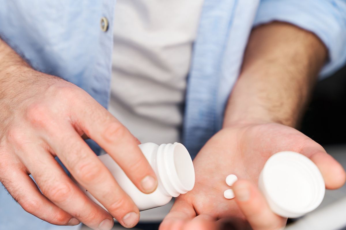Close-up view of man in blue button-down shirt placing a pill in his hand. Man taking two pain pills.
