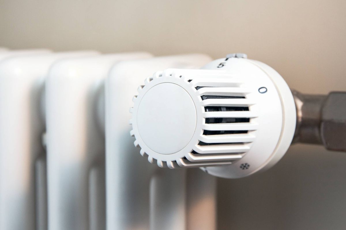 An image displaying a close-up view of a white thermostat with a circular dial, attached to a central heating radiator. The radiator features multiple vertical slats and is mounted on a beige wall.