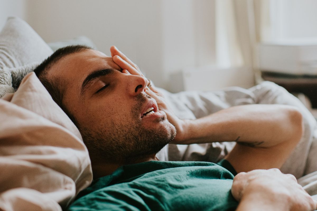 A man has just woken up in a domestic environment, either a living room or a bedroom. He is cozy in duvets and pillows. He rubs his eyes and face sleepily as he comes to.