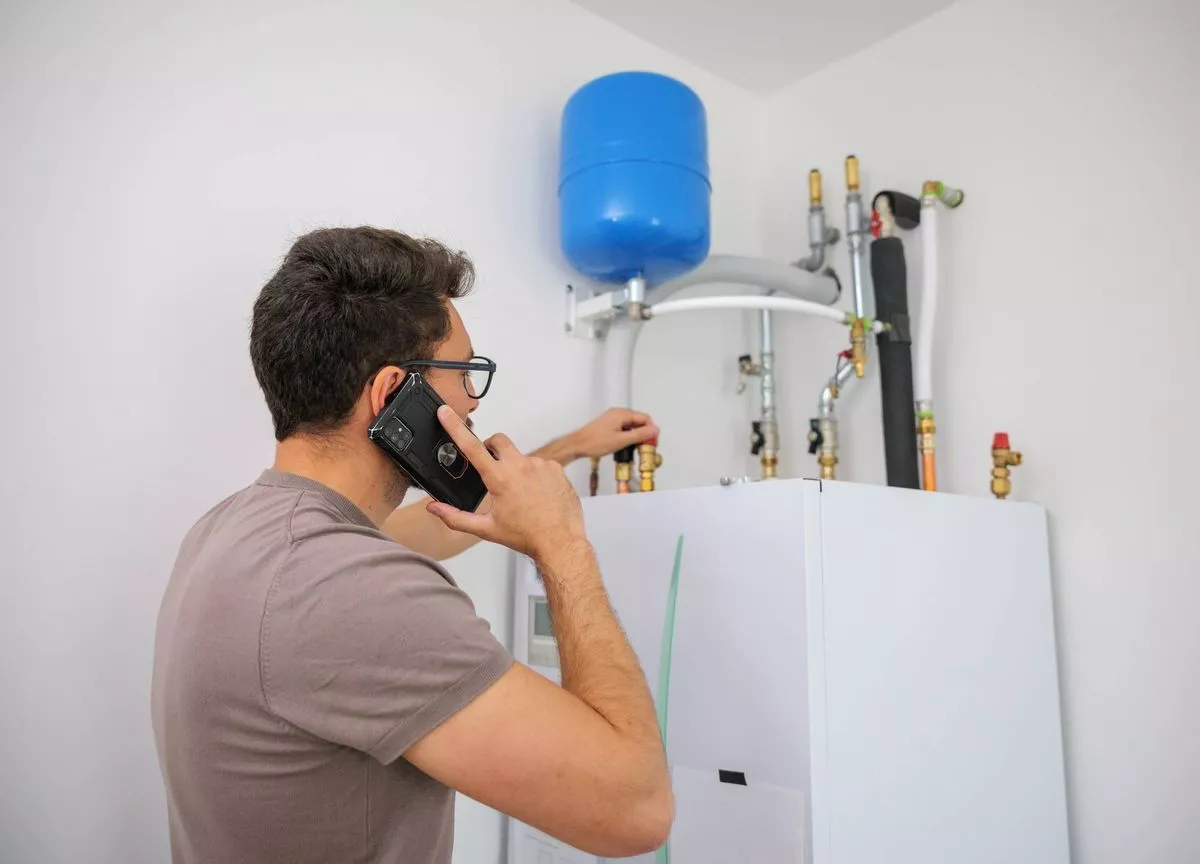 An individual in a casual attire, engaged in a conversation on a mobile phone, standing beside a water heater apparatus. The water heater is a white cylindrical unit, with visible pipes and a blue tank positioned above it. The setting appears to be an indoor environment with a plain white wall in the background.