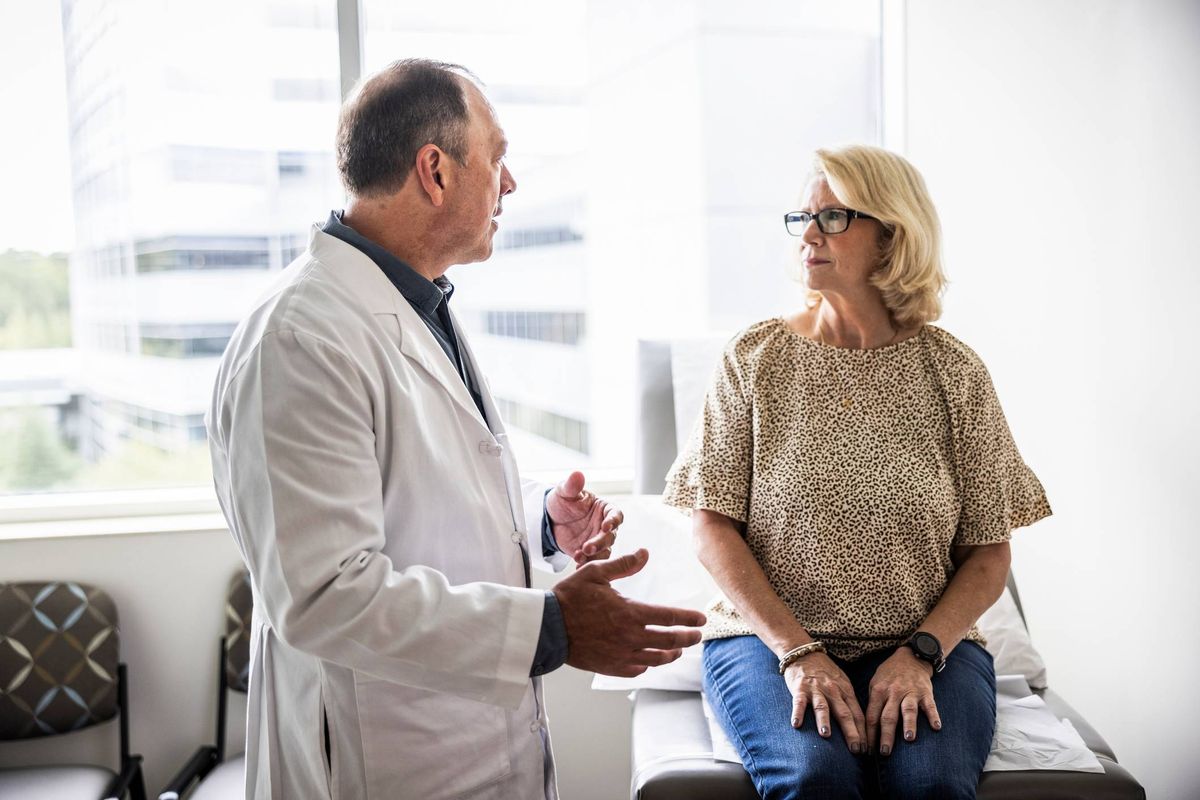 A healthcare professional in a white lab coat is engaged in a discussion with a female patient, who is seated and attentively listening. The setting appears to be a modern medical examination room.