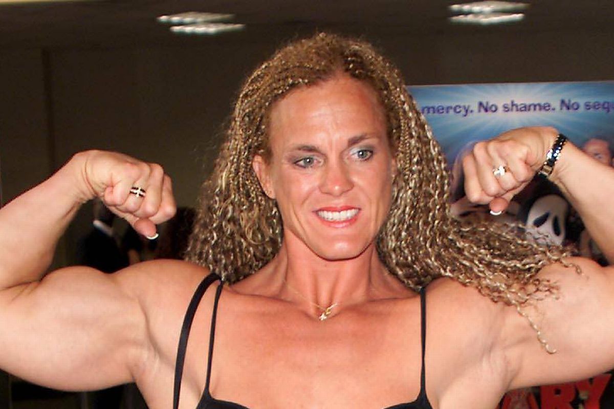 An adult female, dressed in a black cut-out dress, flexes her muscular arms in a room adorned with various posters in the background.