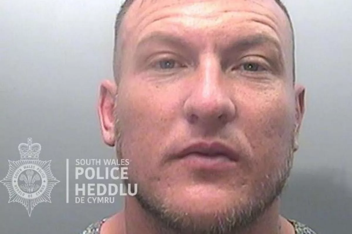 A police mugshot of a white man in his late 30s. He has a light beard and short hair and appears to have a tattoo to the left side of his neck
