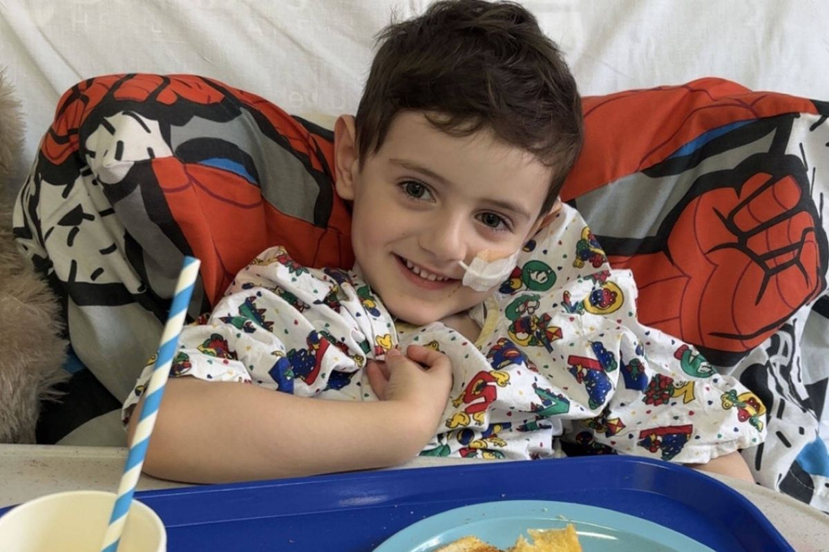 A young child with a cheerful expression lies on a hospital bed, wearing a colorful printed shirt. The child has a tube inserted in their nostril, and they are surrounded by vibrant pillows with a mix of red, gray, and black patterns.