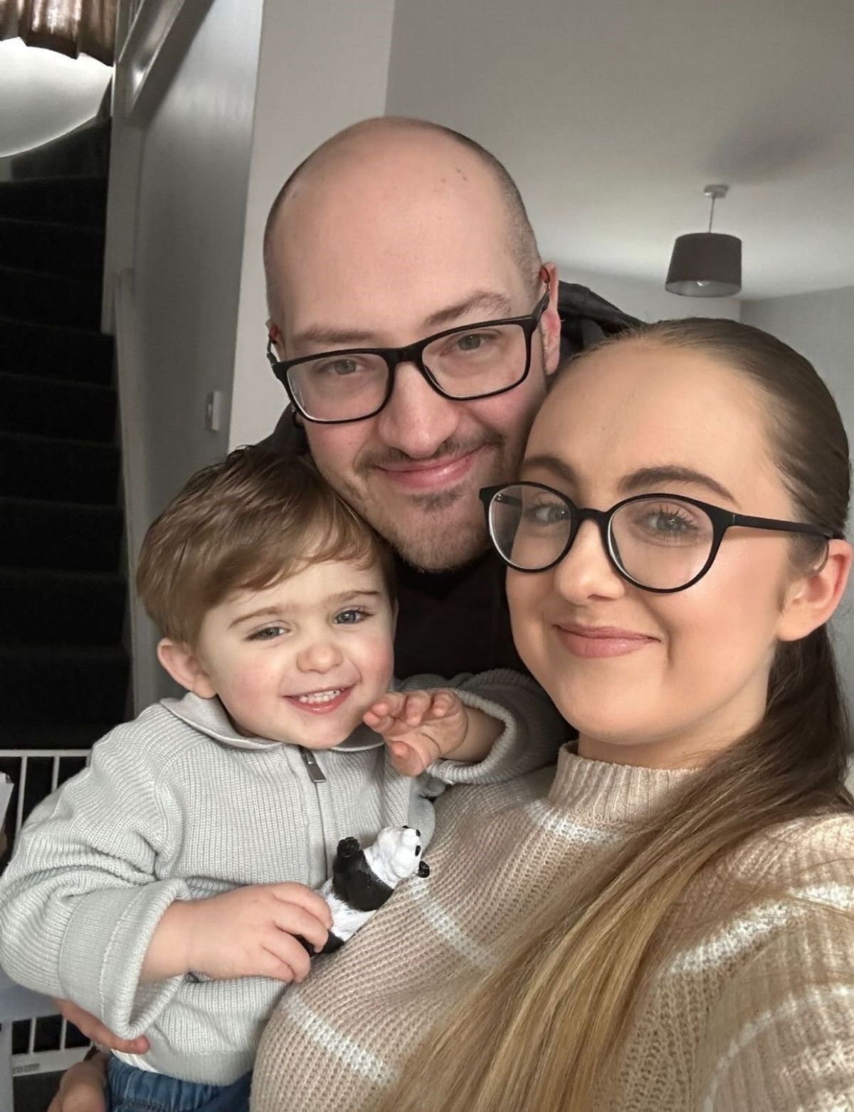 A young woman with glasses, standing next to a bald man with glasses, they are smiling as she holds a three year old boy