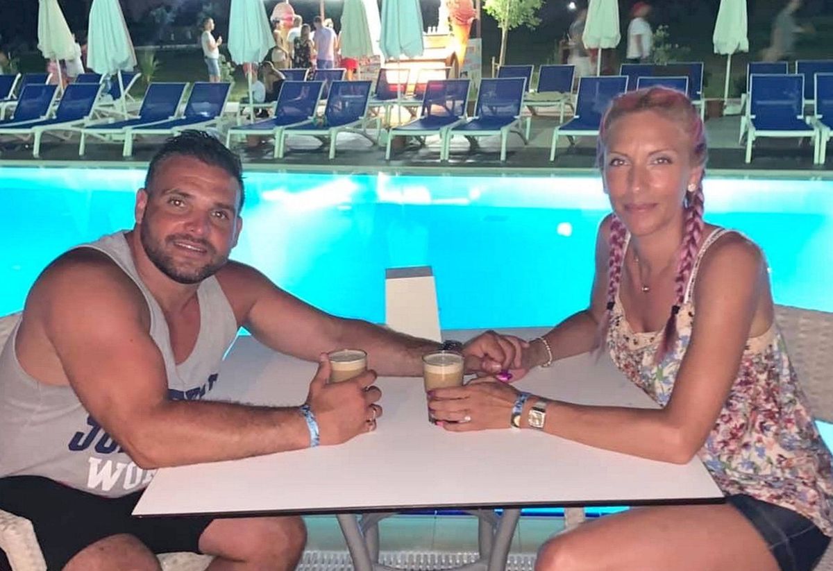 Two individuals, dressed casually, are seated at an outdoor table adjacent to a swimming pool, holding drinks in their hands. The setting includes a row of lounge chairs and umbrellas in the background.