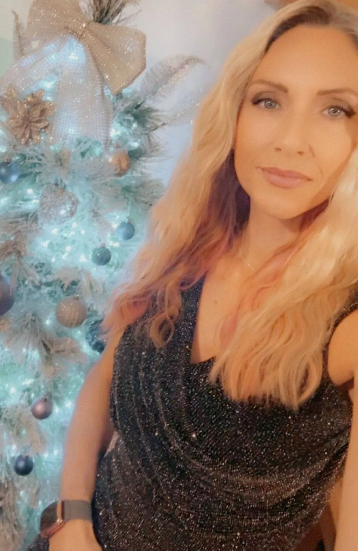 An individual with long blonde hair is standing in front of a decorated Christmas tree, wearing a black sleeveless dress and a wristwatch.