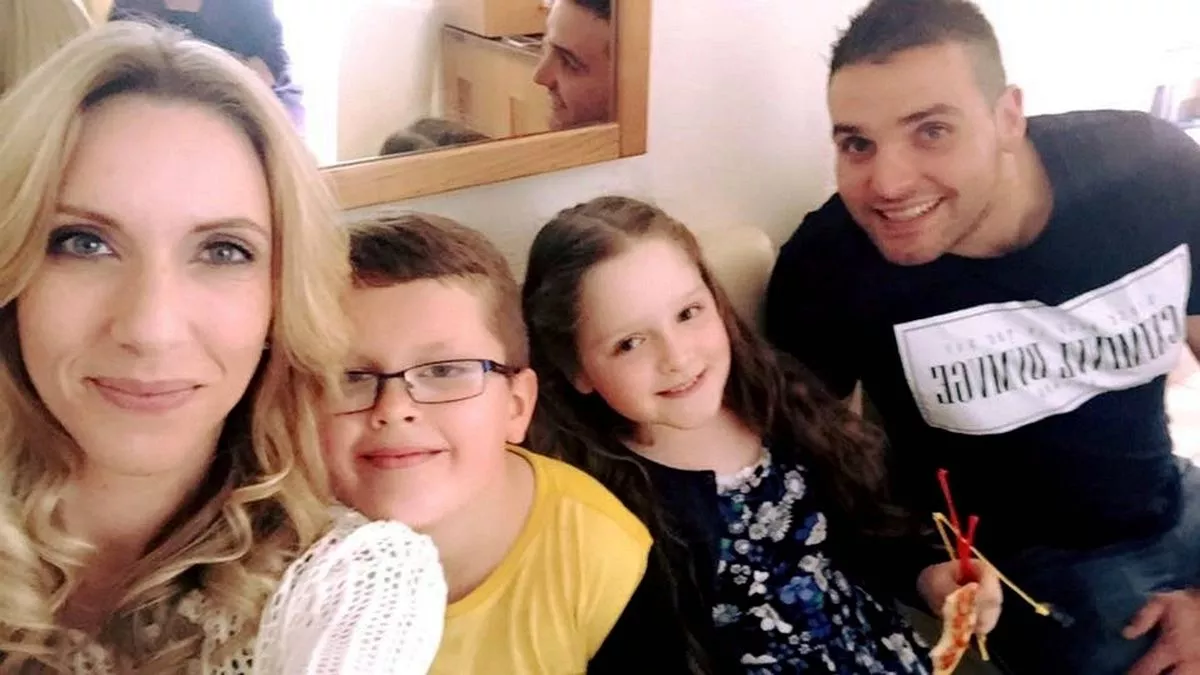 A family of four is seated together on a couch, likely posing for a family photograph. The adult woman, on the left, wears a warm smile and a light-colored top. Next to her is a child with glasses, dressed in a yellow shirt. To the right, a young girl with braided hair is holding a craft item. The adult man, on the far right, wears a black t-shirt with white text. Behind them, a wall adorned with a framed picture is visible.