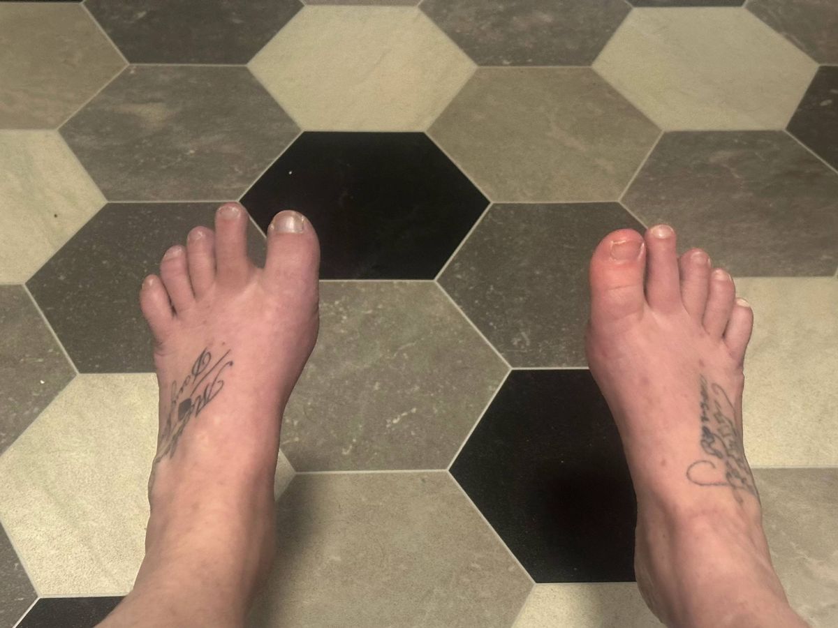 A pair of bare feet positioned on a tiled floor featuring a hexagonal pattern in varying shades of beige and black. The floor tiles display a symmetrical arrangement, creating a geometric design. The individual's feet exhibit a tattoo on the left foot's arch and a similar design on the right foot's ankle.