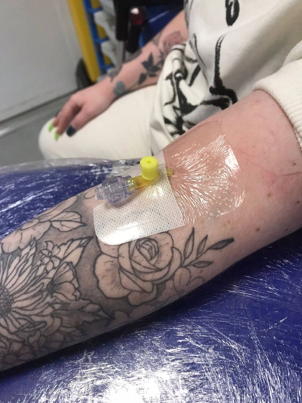 A close-up view of an individual's forearm, showcasing a freshly applied adhesive patch over a partially completed tattoo of a rose. The arm rests on a blue protective surface, while the individual's hand is positioned nearby, partially visible in the background.