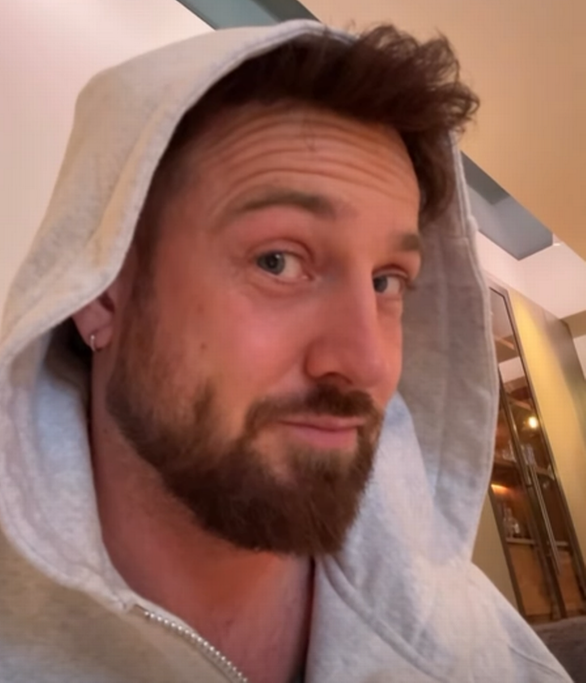 Sam Thompson cheekily raises his eyebrows as he wears a grey hoody with the hood up