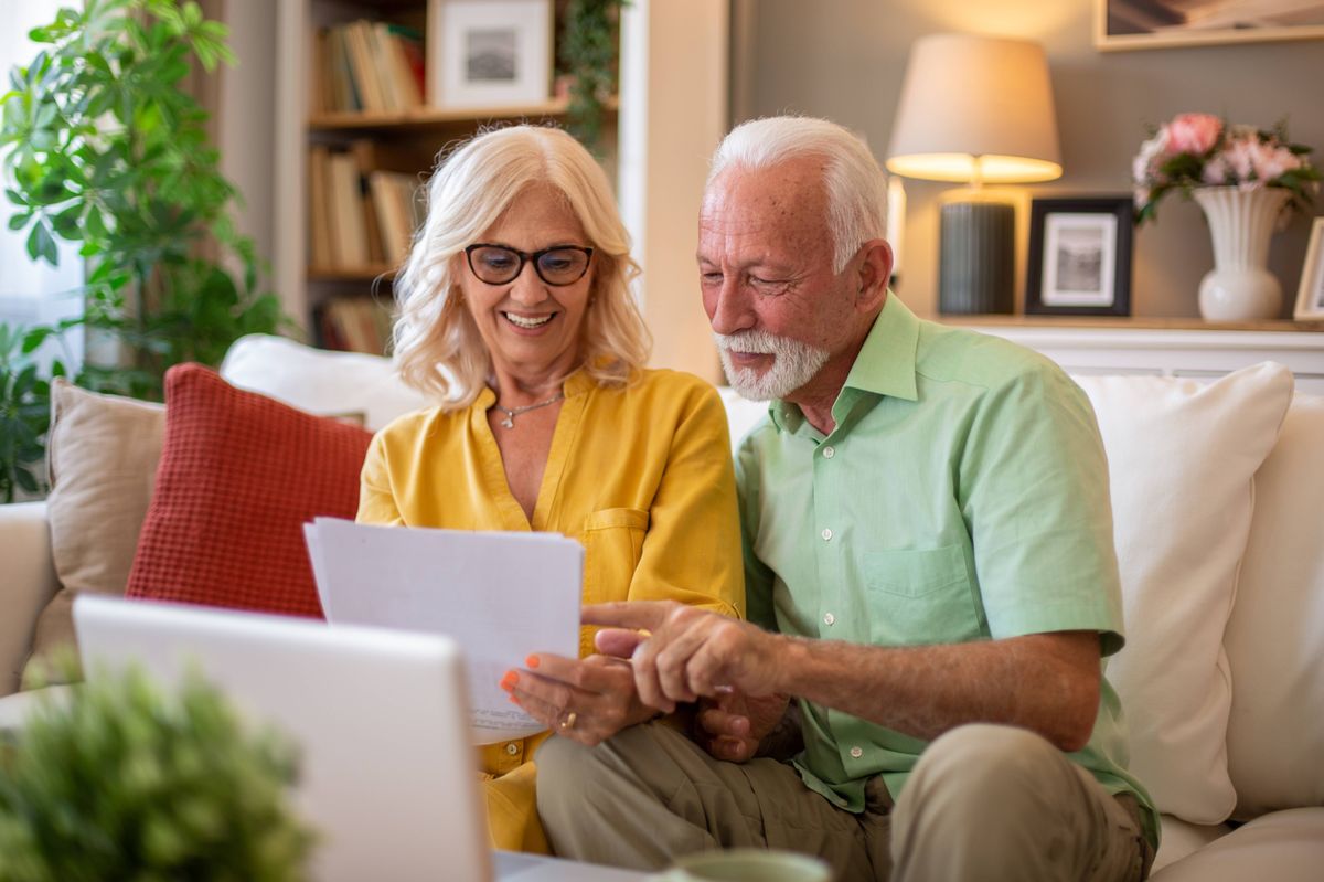 Happy retired couple calculating domestic bills and checking financial documents, using laptop at home