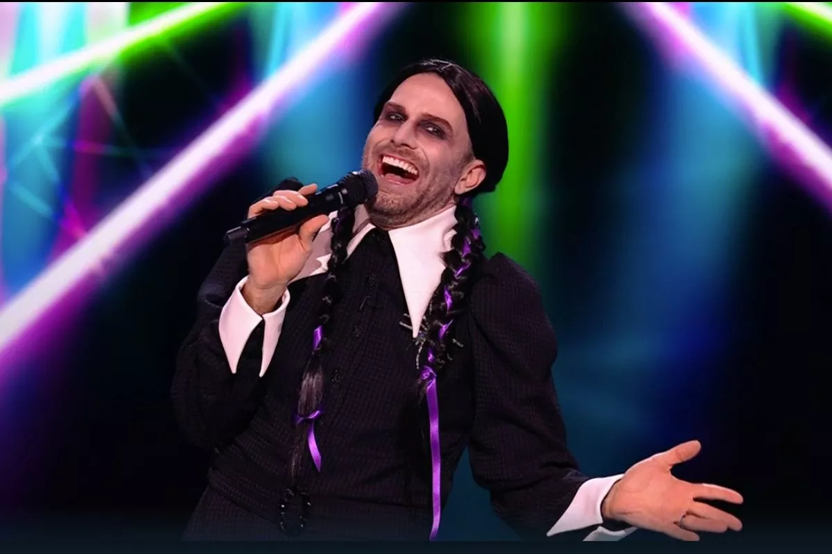 Joel Dommett on tonight's episode dressed as Wednesday Addams in black plaited wig and a black dress with a white collar. He is holding a microphone and smiling 