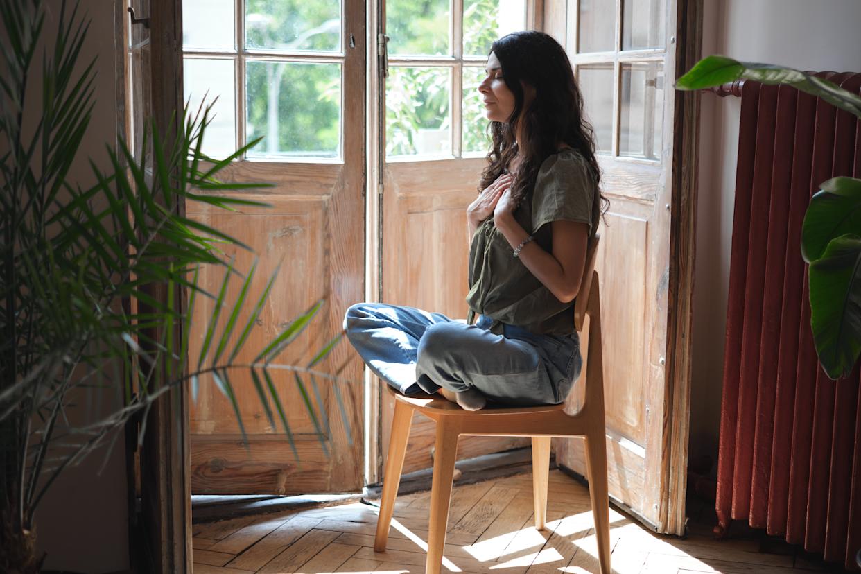 Calm serene woman sitting in lotus pose on chair with closed eyes, hands on chest, practising deep breathing exercise to find peace and mindfulness