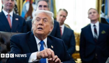 Donald Trump pictured sat behind his desk in the Oval Office. He wears a black suit jacket, white shirt, blue tie, and a pin of the US flag. Behind him stand four male officials in suits.