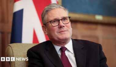 British Prime Minister Sir Keir Starmer, wearing black glasses and a dark suit, looks just away from the camera. A British flag is draped in the background.