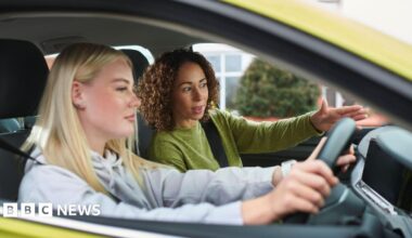 A young girl with long blond hair is behind the wheel of a yellow car. There is a woman beside her, with curly hair and a green jumper, giving her instructions.