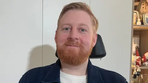 BBC Michael Campbell has a ginger beard and blue eyes.  The top of his wheelchair is visible behind his head. He is wearing a navy shirt with a white T-shirt underneath.