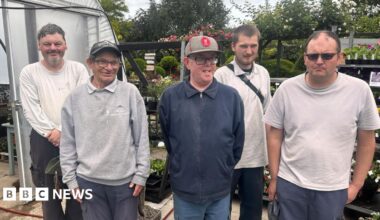 Five men, varying in age, standing in a row in front of stand of plants