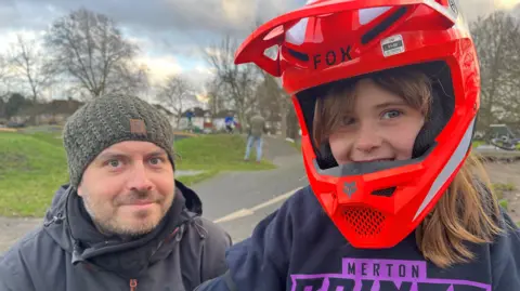 Man with a green hat and dark jacket stands with young girl who wears bright red BMX helmet. She smiles at the camera.