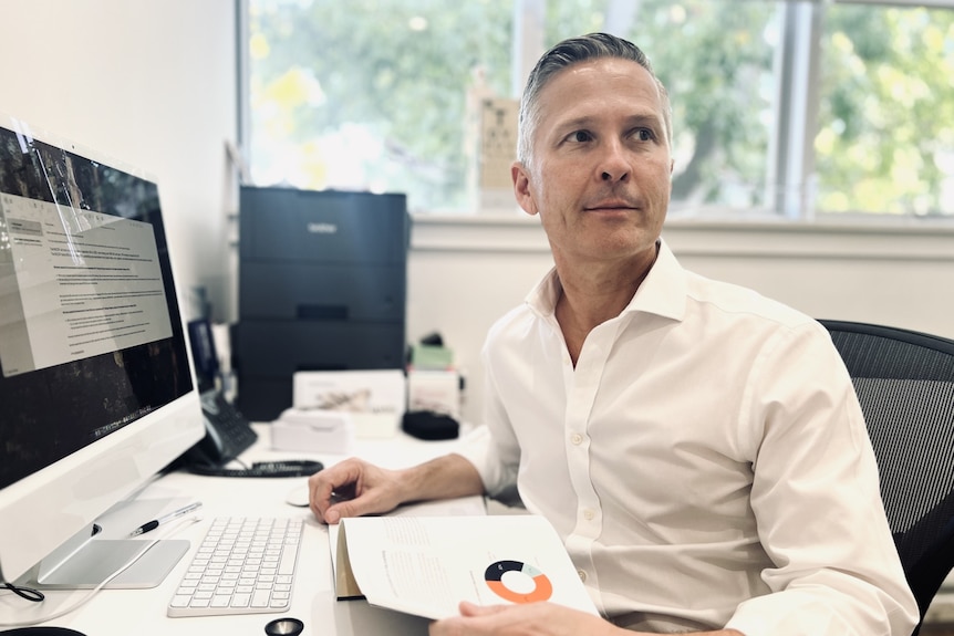 A middle-aged white man with short greying hair and a crisp white shirt sits at a computer in a brightly lit workspace.