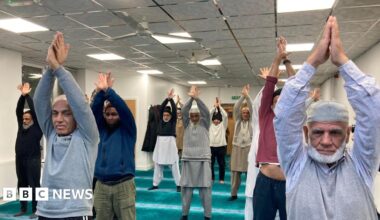 A pilates class is held in a mosque in Bradford. The men participating are in comfortable, casual clothing and traditional dress. All are standing with their arms raised in the air.