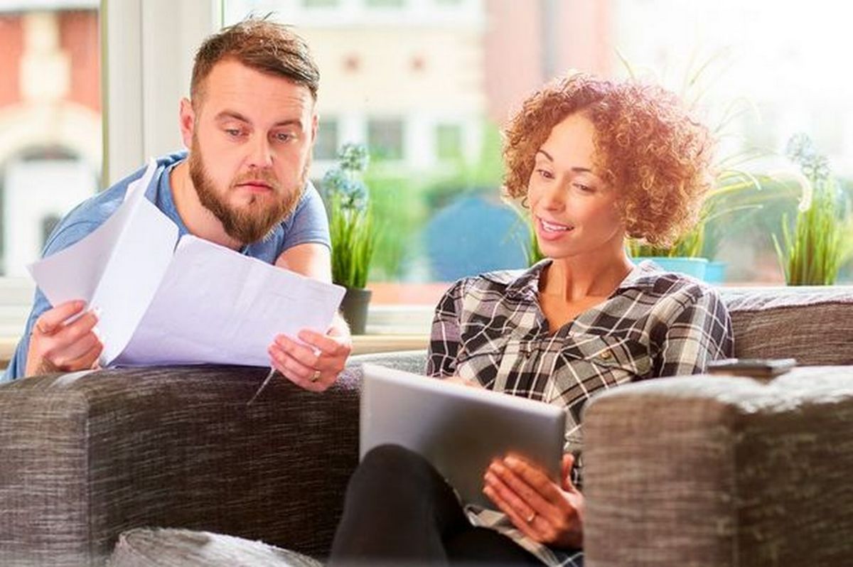 A young man and woman are looking at a computer and printed document with happy expressions on their faces. 