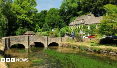 Liam Gallagher wears a black raincoat and sunglasses as he tilts his head upwards to sing into a microphone on stage. Spotlights are shining behind him. He is holding his arm out with his thumb sticking up as he sings.