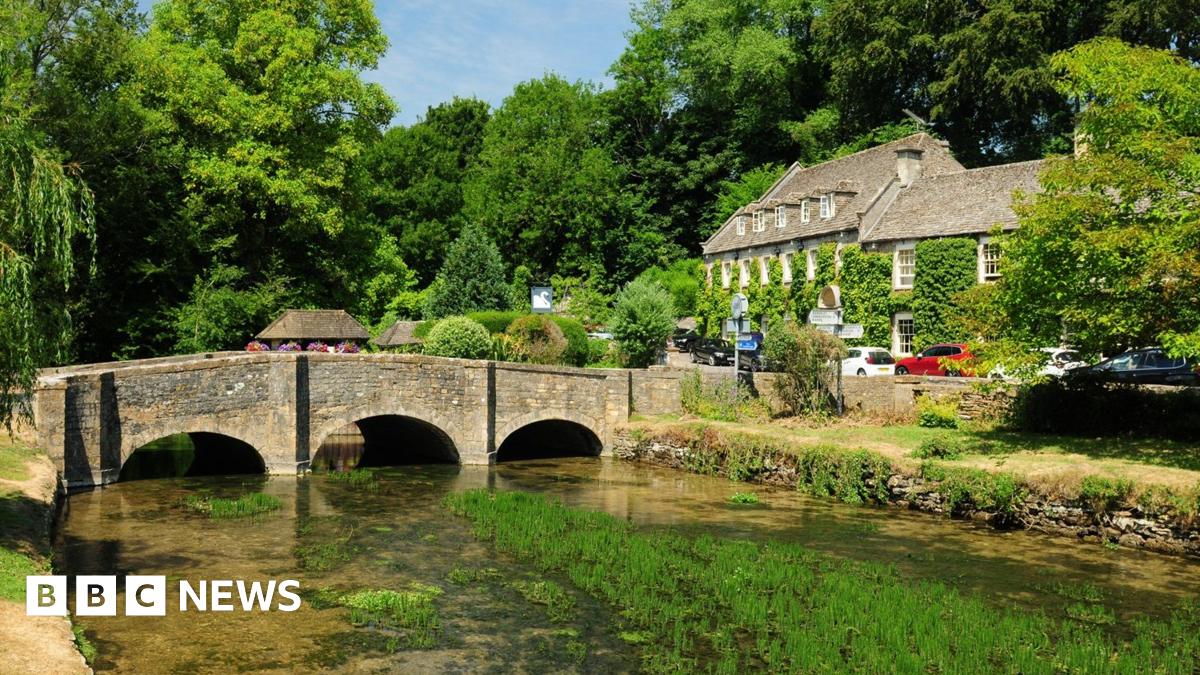 Liam Gallagher wears a black raincoat and sunglasses as he tilts his head upwards to sing into a microphone on stage. Spotlights are shining behind him. He is holding his arm out with his thumb sticking up as he sings.