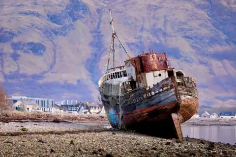 Graham Christie A large, rusted fishing boat lies tilted on a rocky shoreline, its wooden hull weathered and broken. Behind it, a small coastal town sits beneath steep, rugged hills under a pale sky.