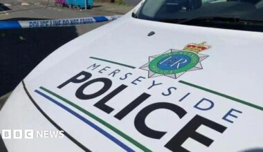 The white bonnet of a Merseyside Police patrol car, bearing the name of the force in black and blue lettering. Police tape can be seen across a road behind the vehicle.