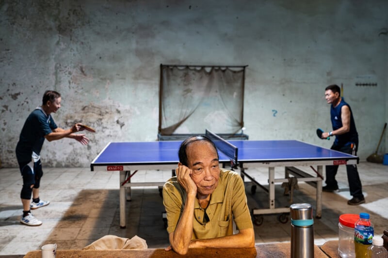 An elderly man sits at a table with his head resting on his hand, looking thoughtful, while two men play table tennis in the background in a worn, dimly lit room.