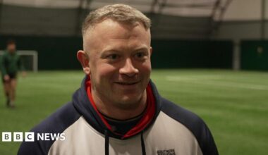 Dan Robinson is  a white man, wearing a white hoody, with blue sleeves and good, he has cropped grey hair and is smiling. He's standing on a green all-weather pitch where some of his fellow rugby players can be seen warming-up in the background.