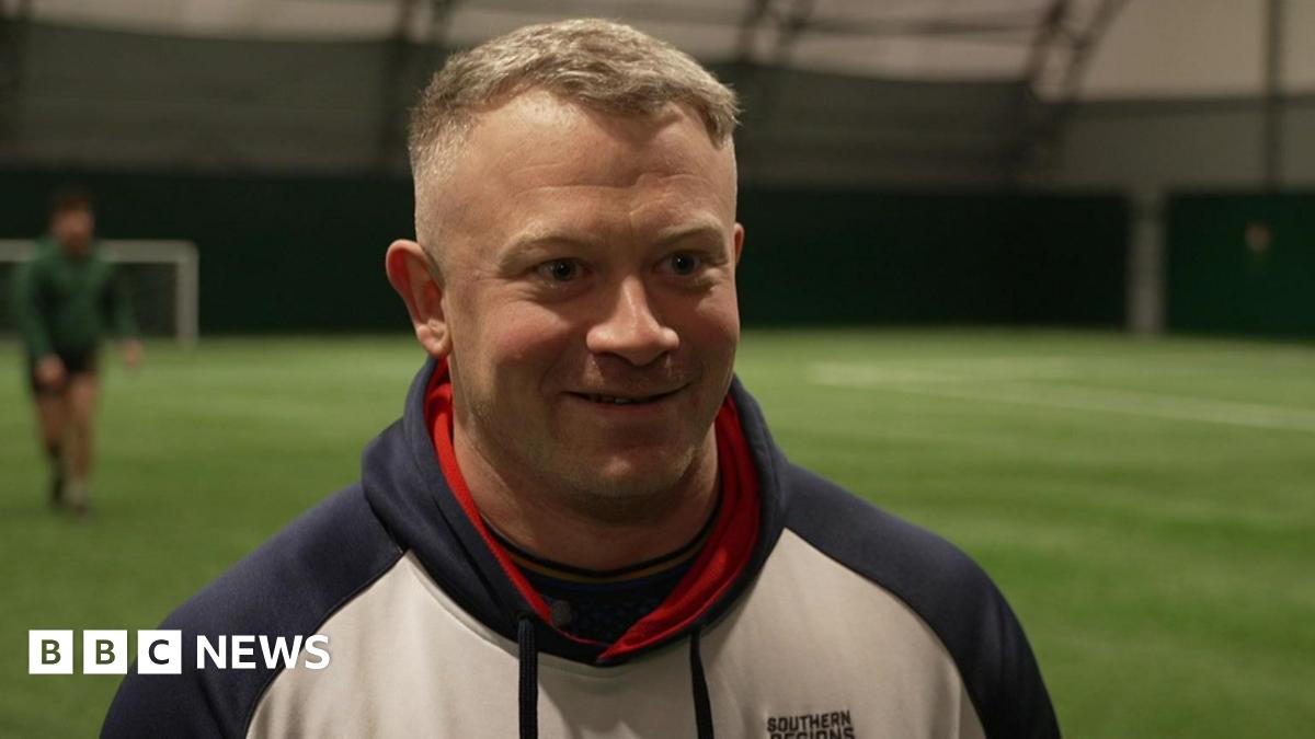 Dan Robinson is  a white man, wearing a white hoody, with blue sleeves and good, he has cropped grey hair and is smiling. He's standing on a green all-weather pitch where some of his fellow rugby players can be seen warming-up in the background.