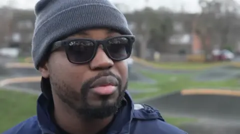 Man in sunglasses with grey hat looks away from camera. In the background is the BMX park.