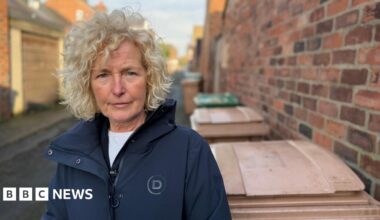 A woman with blonde curly hair is wearing a dark blue coat. She is standing in a alleyway beside brown wheelie bins and a high red-brick wall.