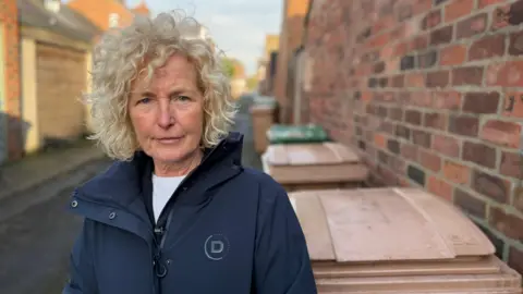 Becki Bowden / BBC A woman with blonde curly hair is wearing a dark blue coat. She is standing in a alleyway beside brown wheelie bins and a high red-brick wall.