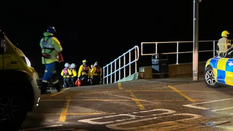 Amanda White/BBC Lifeboat crews on a ramp to the beach area at Withernsea. They are dressed in yellow and black and are wearing helmets. There is a parked police car in the foreground.