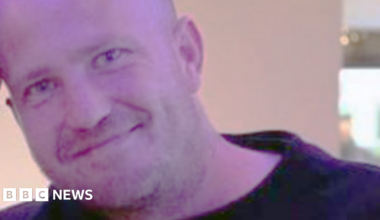 Derek Russell, a shaven headed man wearing a black top, smiles for the camera while standing indoors.