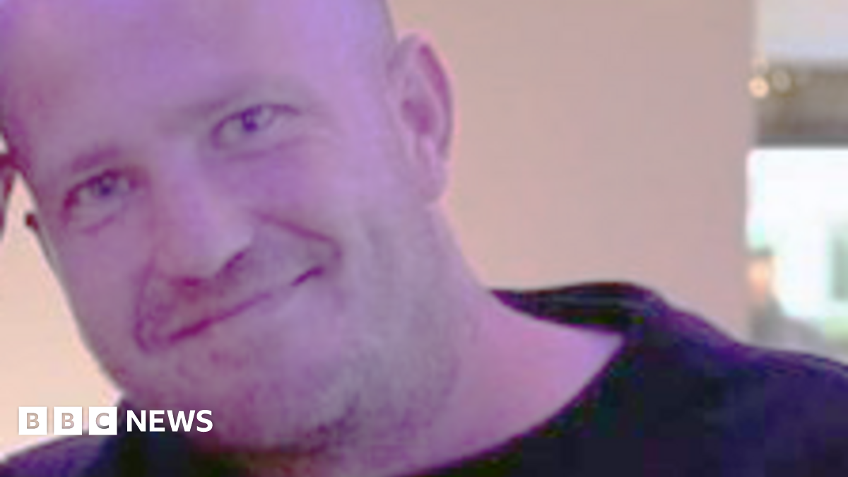 Derek Russell, a shaven headed man wearing a black top, smiles for the camera while standing indoors.