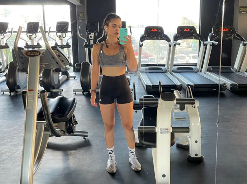 A fit woman in form-fitting shorts and a crop top takes a full-body selfie of herself in the mirrors that also show fitness machines behind her. 