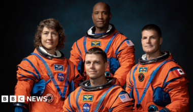 The image shows four astronauts in bright orange space suits posing for a formal group portrait against a dark background. The suits are bulky with blue joints, straps and pockets, emphasising their technical design. Three astronauts stand behind while one is seated or crouching in front, all facing the camera. Their faces are blurred into smooth ovals so no expressions can be seen, keeping the focus on the uniforms. Mission-style patches and flags, including US and Canadian flags, are visible on their arms and chests. Soft, focused lighting makes the vivid orange suits stand out dramatically.