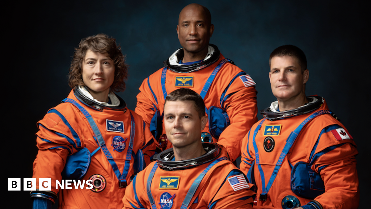 The image shows four astronauts in bright orange space suits posing for a formal group portrait against a dark background. The suits are bulky with blue joints, straps and pockets, emphasising their technical design. Three astronauts stand behind while one is seated or crouching in front, all facing the camera. Their faces are blurred into smooth ovals so no expressions can be seen, keeping the focus on the uniforms. Mission-style patches and flags, including US and Canadian flags, are visible on their arms and chests. Soft, focused lighting makes the vivid orange suits stand out dramatically.