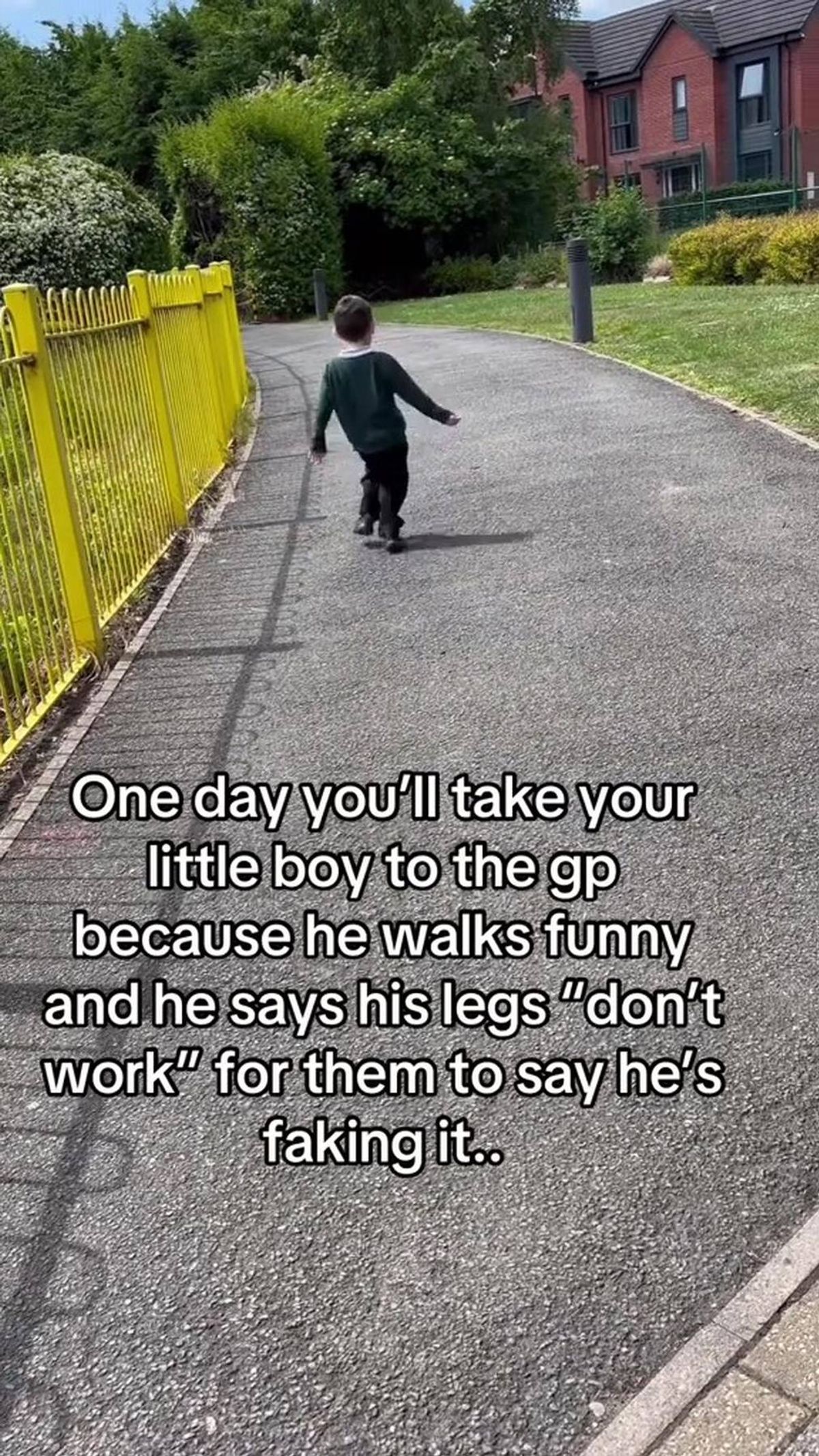 A child in a dark outfit is running along a paved pathway, adjacent to a yellow metal fence. The surrounding area includes lush green grass and shrubs, with the sun casting shadows on the ground.