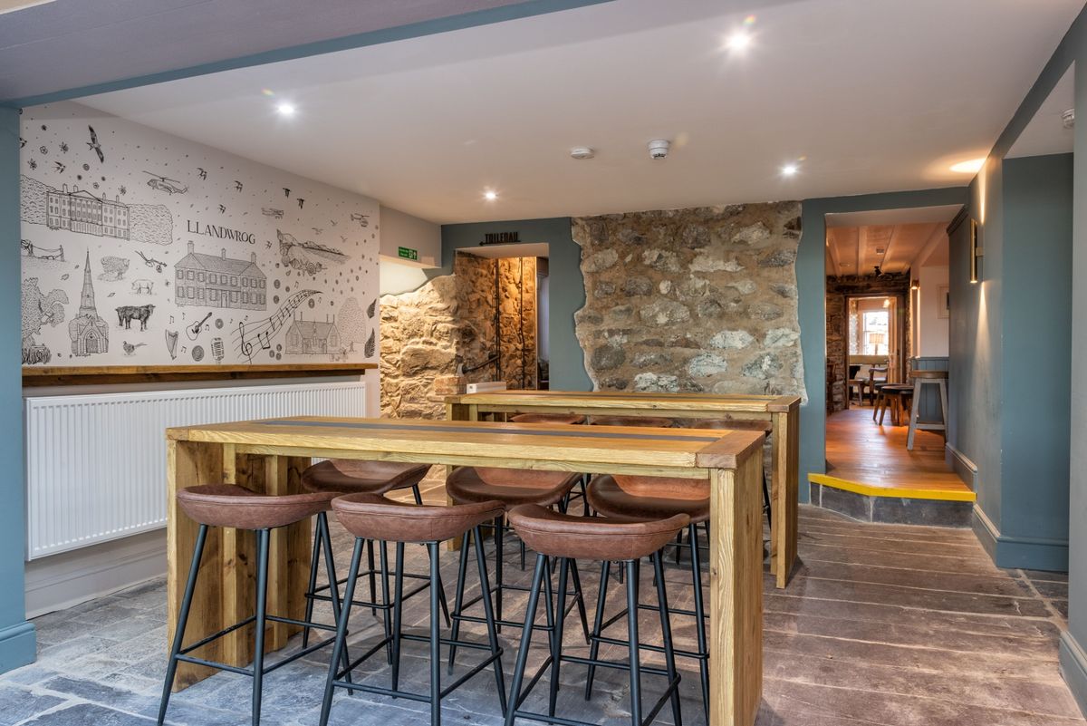 The transition from old to new is this light-filled space that features original exposed stone walls and a village wall mural  at the renovation, conversion and transformation of T'yn Llan commnity pub in Llandwrog, Gwynedd this is the new build extension with restaurant, catering kitchen, facilities and community room